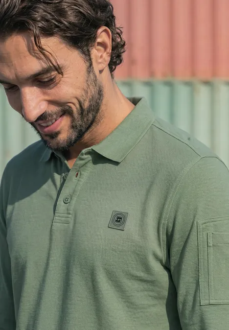 Poloshirt met lange mouwen en zakdetail willow green Een lachende man met een groen poloshirt kijkt omlaag voor een pastelgekleurde achtergrond.
