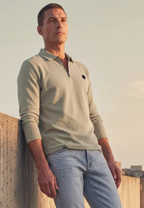 Poloshirt met lange mouwen en ritsdetail antique beige Een man leunt tegen een muurtje, draagt een licht polohemd en spijkerbroek en kijkt in de verte bij zonsondergang.