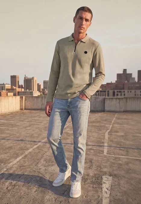 Poloshirt met lange mouwen en ritsdetail antique beige Een man op een dak met een lichtgroen poloshirt, spijkerbroek en witte sneakers tegen een stadsachtergrond.