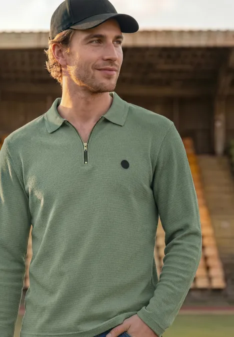 Poloshirt met lange mouwen en ritsdetail willow green Een man met groen langarm-polo en zwarte pet staand voor tribunes.