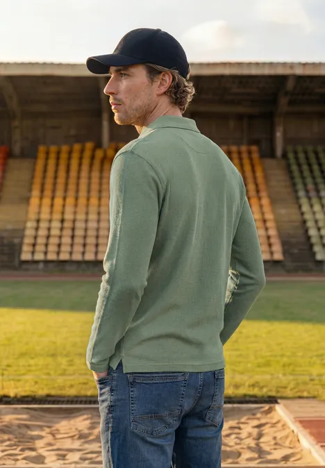 Poloshirt met lange mouwen en ritsdetail willow green Een man in groen langarm-polo en pet, zijwaarts, voor lege tribunes.