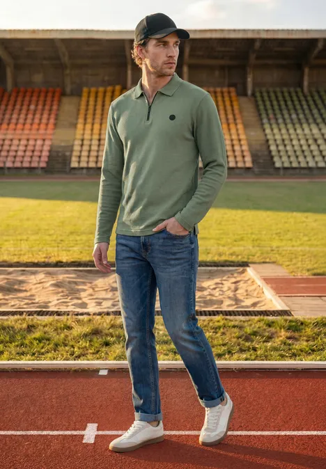 Poloshirt met lange mouwen en ritsdetail willow green Een jonge man in olijfgroen poloshirt, spijkerbroek en pet staat op de atletiekbaan voor de tribunes.