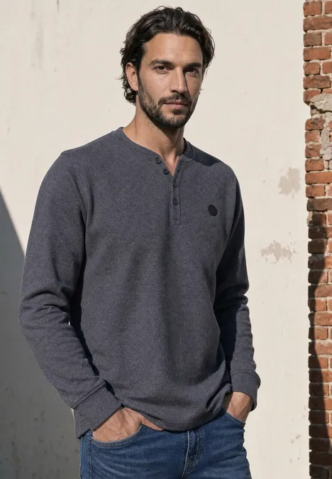 Ein Mann lehnt an einer Mauer und trägt ein dunkelblaues Henley-Shirt.