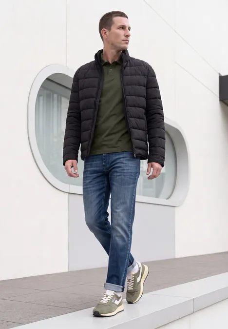 Un homme en veste matelassée foncée, polo vert, jean et baskets marche devant un bâtiment moderne.