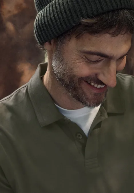 Un homme souriant portant un bonnet et une barbe naissante regarde vers le bas sur un arrière-plan flou et chaleureux.