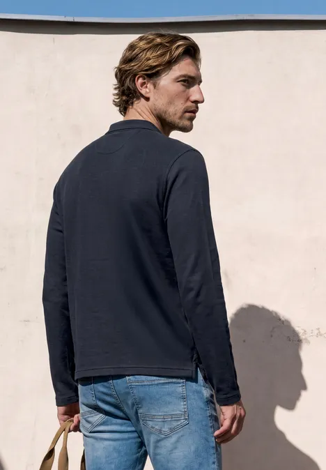 Langarm Poloshirt in Unifarbe dark ink blue Ein Mann mit blonden Haaren steht vor einer pastellfarbenen Wand, trägt dunkles Oberteil und Jeans.