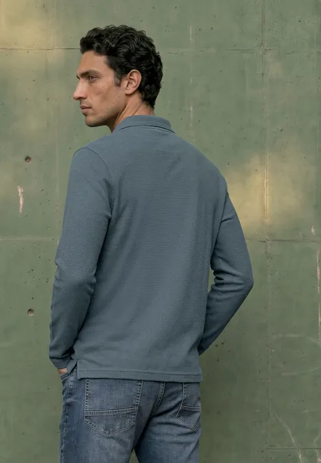 Langarm Poloshirt in Unifarbe vintage blue Ein Mann in blauem Polohemd und Jeans steht vor einer grünen Wand und blickt nach rechts.