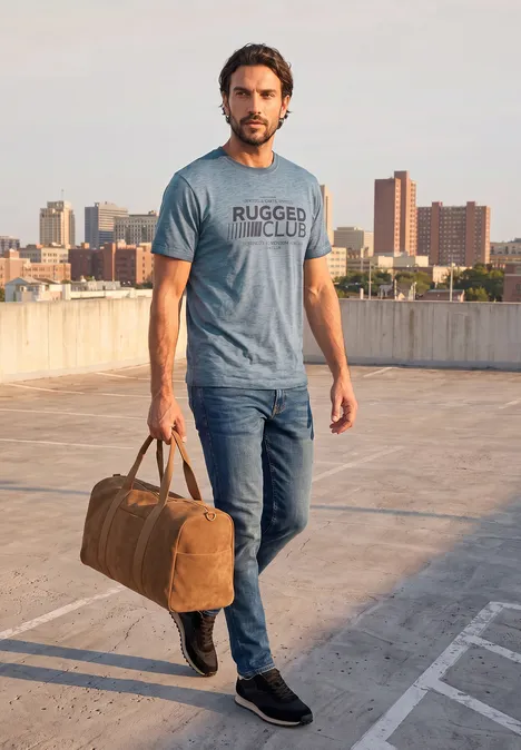 Ein Mann in Jeans und Shirt trägt eine Reisetasche und steht auf einem Dach mit Stadtkulisse im Hintergrund.
