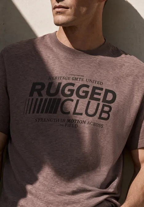 Mann in braunem T-Shirt mit schwarzem 'RUGGED CLUB'-Schriftzug vor hellem Hintergrund.