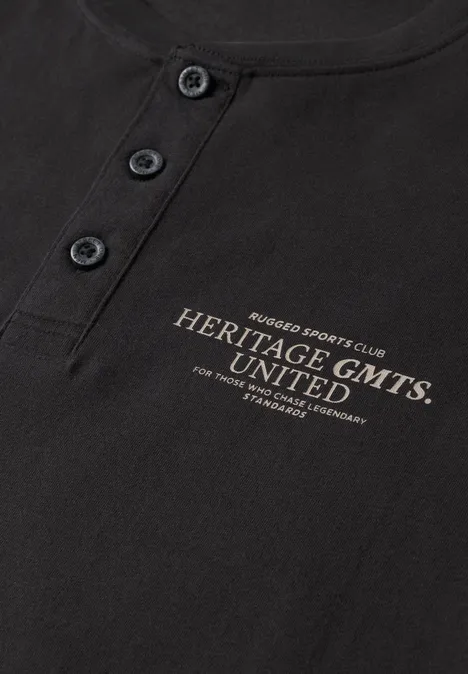 T-shirt noir avec patte de boutonnage et inscription dorée 'Heritage GMTS UNITED'.
