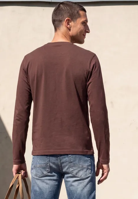Een man van achteren in een bruine longsleeve en spijkerbroek, met een gele tas voor een lichte muur.