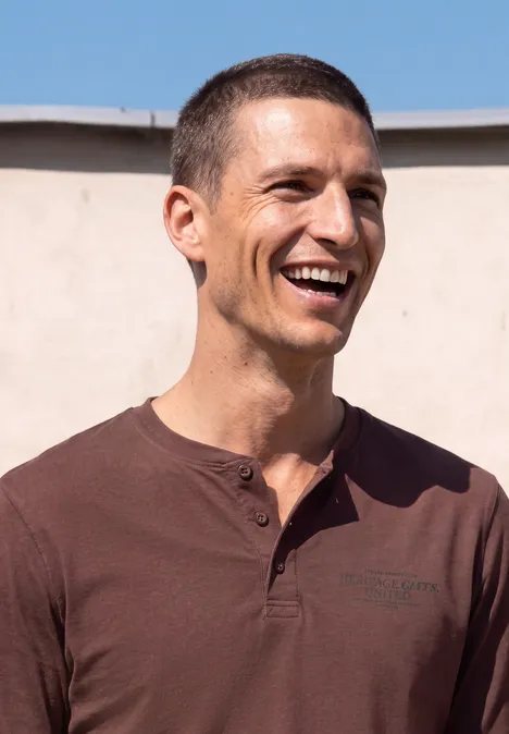 Een lachende man met kort donker haar en bruin shirt voor een lichte muur.