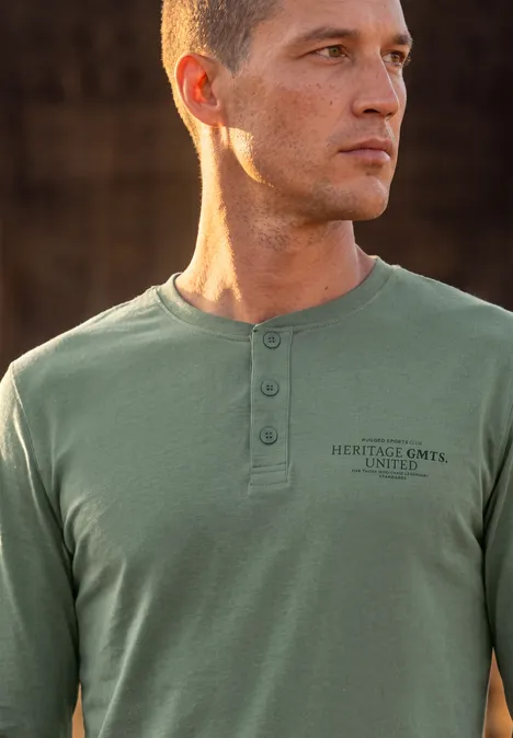 Un jeune homme aux cheveux courts porte un t-shirt vert olive à manches longues avec patte de boutonnage et inscription.