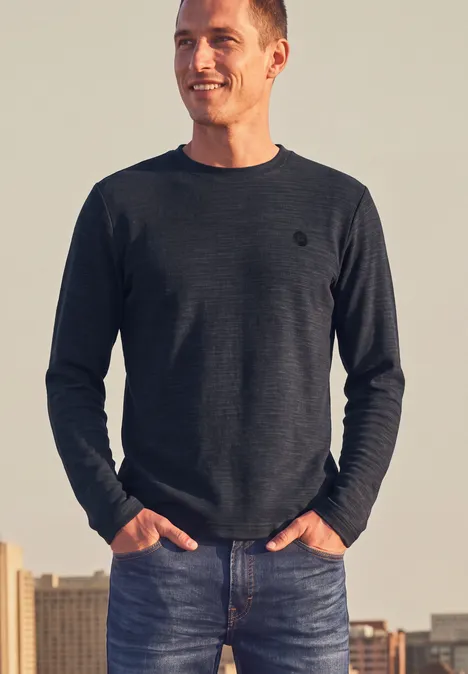 Ein lächelnder Mann in dunkelgrauem Pullover und Jeans steht mit Händen in den Taschen vor einer Stadt-Skyline bei Sonnenuntergang.