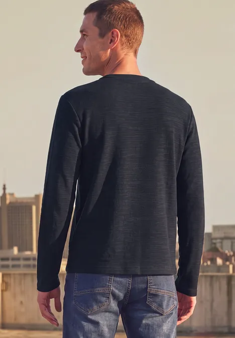 Ein Mann steht auf einem Dach, trägt Jeans und ein dunkles Langarmshirt, Blick zur Stadt im Hintergrund.