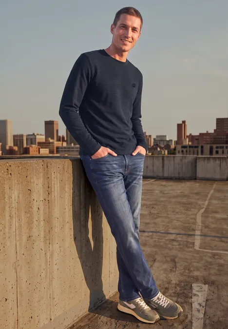 Ein lächelnder Mann in Jeans lehnt auf einer Dachterrasse mit Stadtblick.