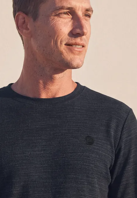 Mann in dunkelgrauem Langarmshirt, schaut seitlich, warmes Licht, dezentes Logo auf der Brust.