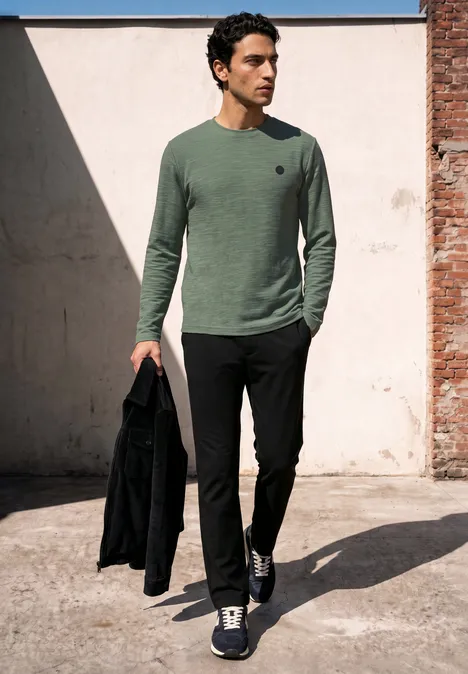 Langarm Shirt mit Rundhals in Unifarbe willow green Ein Mann in grünem Pullover und schwarzer Hose hält eine Jacke und geht draußen in urbaner Umgebung.