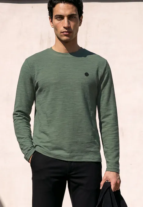 Langarm Shirt mit Rundhals in Unifarbe willow green Ein junger Mann vor einer rosa Wand, trägt ein grünes Langarmshirt, schwarze Hose und hält eine schwarze Tasche.