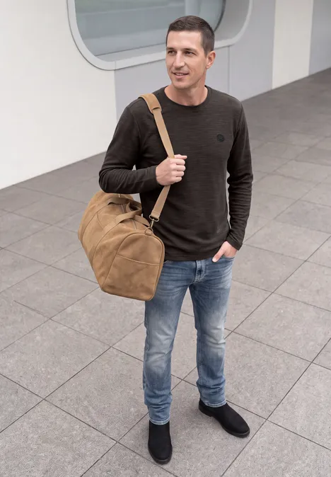 Ein Mann in Jeans und Pullover trägt eine Reisetasche und steht in einer Bahnhofsumgebung.