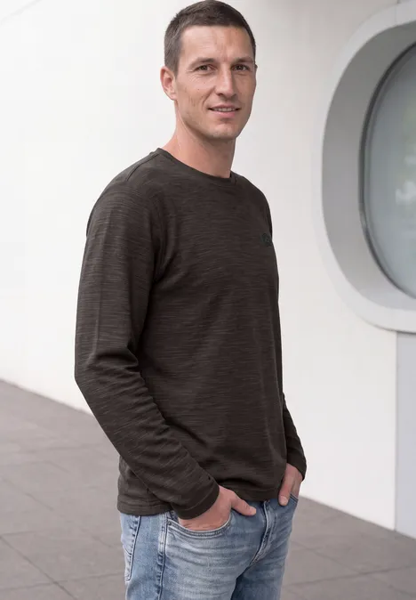 Ein lächelnder Mann in dunklem Pullover und Jeans steht vor einer weißen Wand mit rundem Fenster.