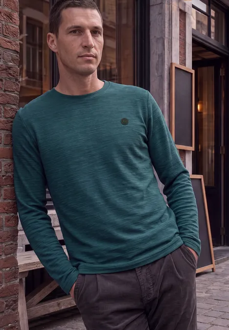 Ein Mann lehnt lässig an einer Backsteinwand, trägt grünes Langarmshirt und graue Hose.