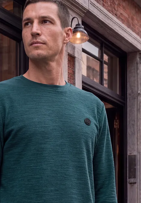 Mann in grünem Langarmshirt mit rundem Logo vor historischer Backsteinfassade.