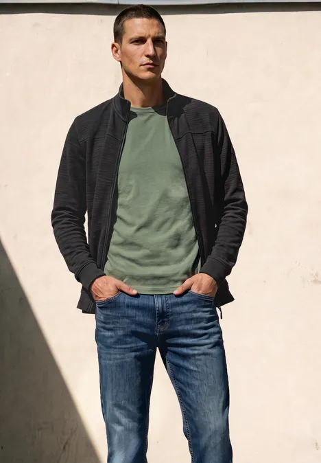 Langarm Jacke mit Stehkragen basic black Ein Mann in grünem T-Shirt, schwarzer Jacke und Jeans steht entspannt vor einer hellen Wand im Sonnenlicht.