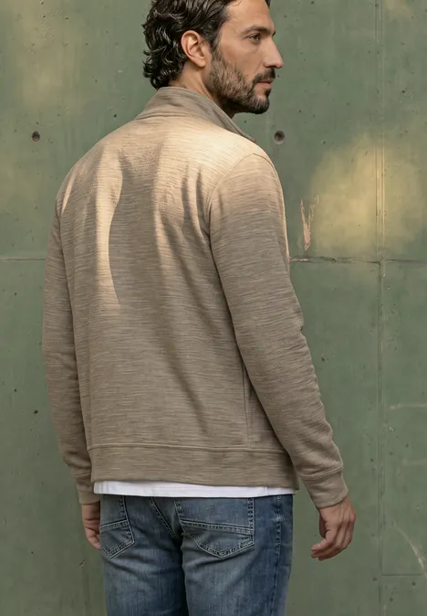 Een man gezien van achteren draagt een beige vest, wit T-shirt en jeans voor een groene muur.