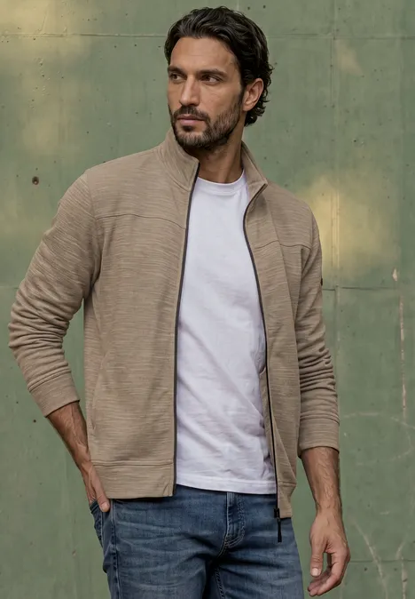 Een man in een beige vest, wit t-shirt en spijkerbroek voor een groene muur.