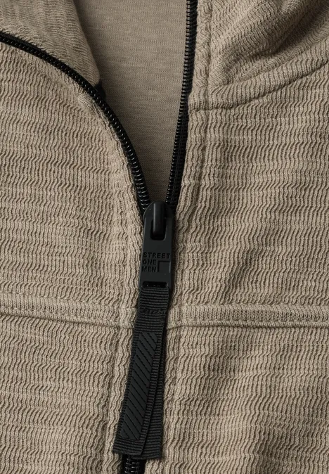 Close-up van een beige textuurvest met rits en zwart ritslipje.