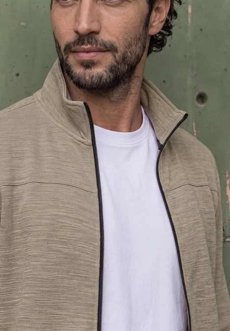Een man met donker haar en baard draagt een beige jas over een wit T-shirt voor een groene achtergrond.