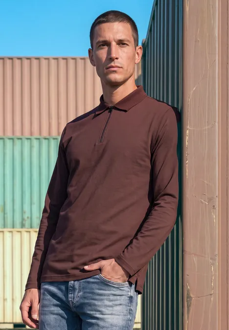 Ein Mann in braunem Poloshirt und Jeans lehnt vor bunten Containern und blauem Himmel.