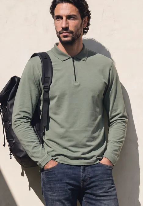 Ein Mann in olivgrünem Langarm-Poloshirt und Jeans mit Rucksack lehnt entspannt an einer hellen Wand.