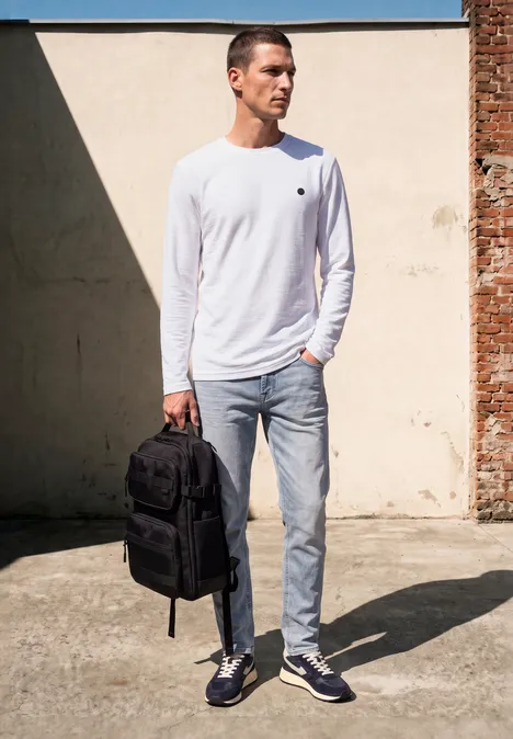 Un homme en t-shirt blanc manches longues et jean porte un sac à dos noir devant un mur clair.