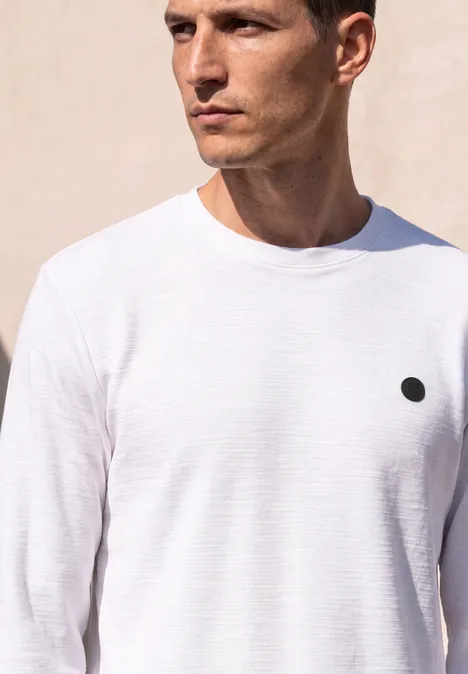Un homme porte un T-shirt blanc simple avec un petit point noir sur la poitrine.