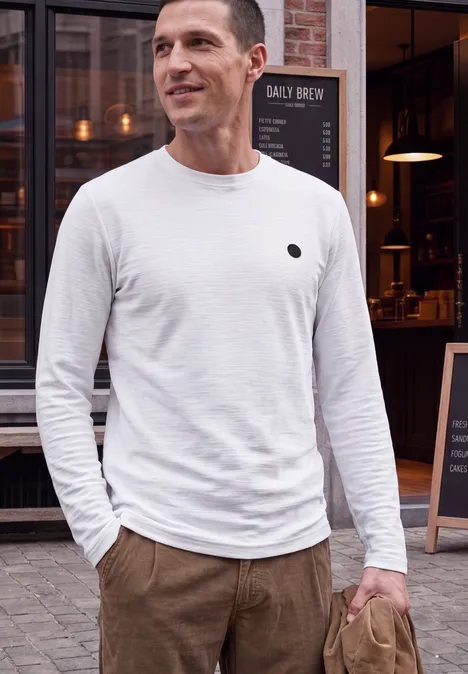 Een man in een wit lang mouw T-shirt en een bruine broek staat voor een café en glimlacht.