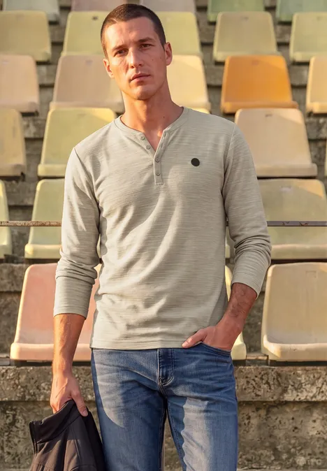 Ein Mann steht vor leeren Stadionrängen, trägt ein graues Langarmshirt, blaue Jeans und hält eine Jacke.