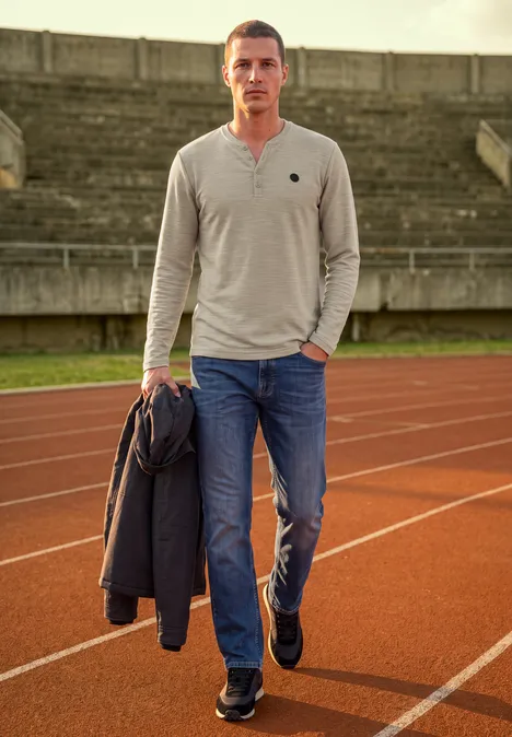 Ein Mann in Jeans und Shirt steht auf einer Laufbahn, er hält eine Jacke in der Hand.