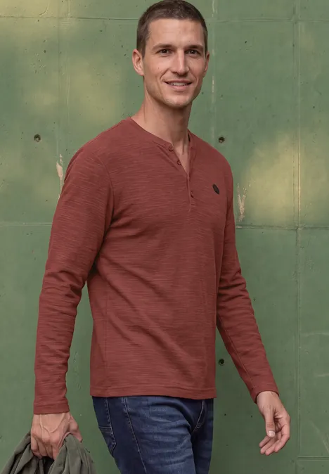Ein Mann in rotem Langarmshirt und Jeans steht vor einer grünen Wand und hält etwas in der Hand.