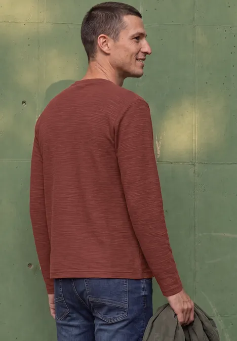Ein Mann mit kurzem Haar steht seitlich, trägt ein rotbraunes Langarmshirt und Jeans und hält eine Jacke.