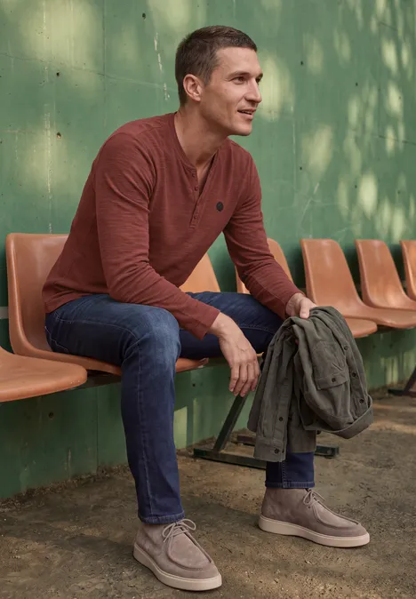 Ein Mann in braunem Pullover und Jeans sitzt auf orangefarbenen Sitzen vor grüner Wand und hält eine Jacke.