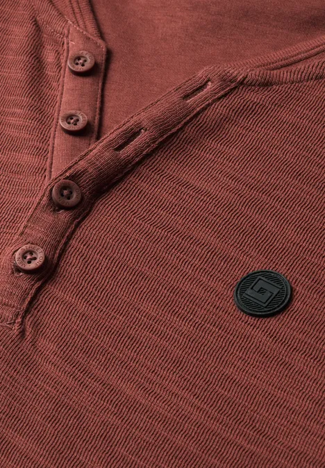 Nahaufnahme eines rostfarbenen Henley-Shirts mit Knopfleiste und rundem, schwarzem Logo-Patch.