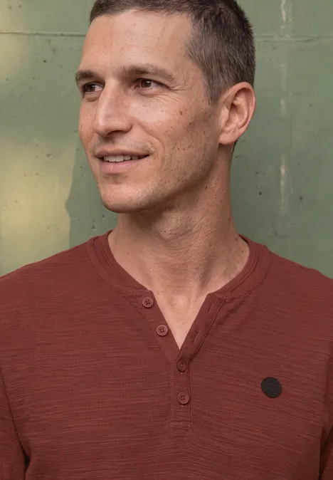Ein lächelnder Mann mit rotem Henley-Shirt vor grünem Hintergrund.