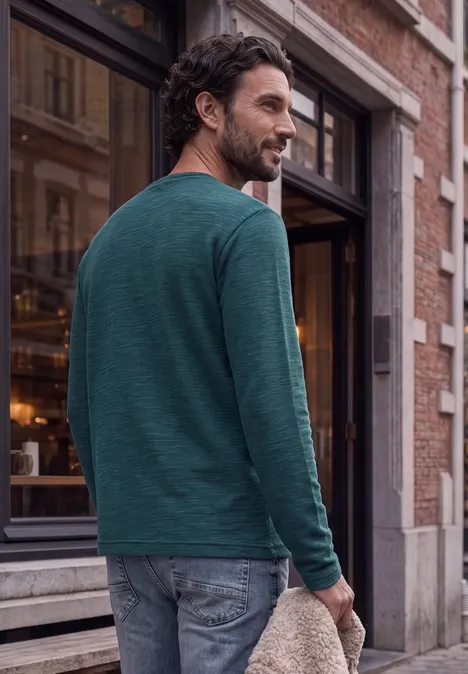 Ein Mann in grünem Langarmshirt und Jeans geht an einem Gebäude vorbei und blickt lächelnd über die Schulter.