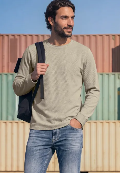 Langarm Shirt mit Popcorn-Struktur classic beige Mann in olivgrünem Pullover, Jeans und Rucksack vor bunten Frachtcontainern.