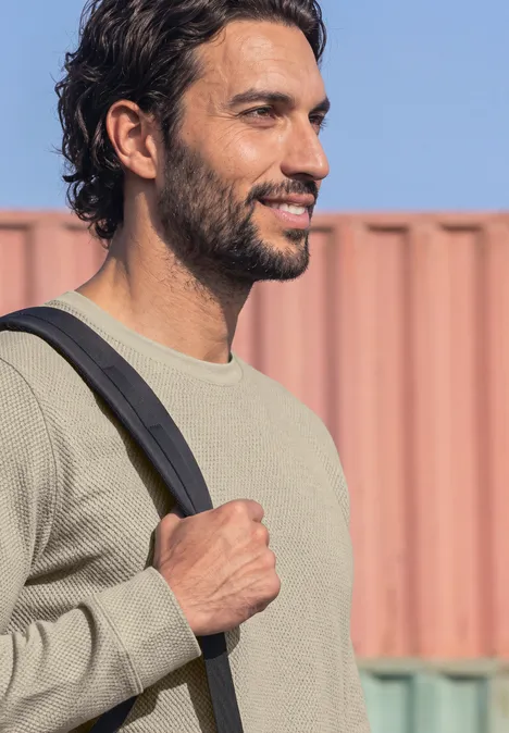 Langarm Shirt mit Popcorn-Struktur classic beige Ein lächelnder junger Mann mit dunklen Haaren trägt seinen Rucksack vor buntem Containerhintergrund.