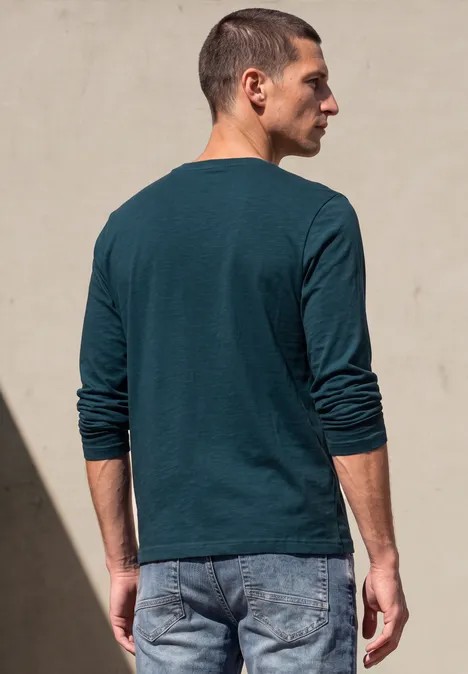 T-shirt à manches longues avec imprimé ton sur ton dark fjord blue Homme vu de dos, en t-shirt vert à manches longues et jean, adossé à un mur clair, éclairé par le soleil.