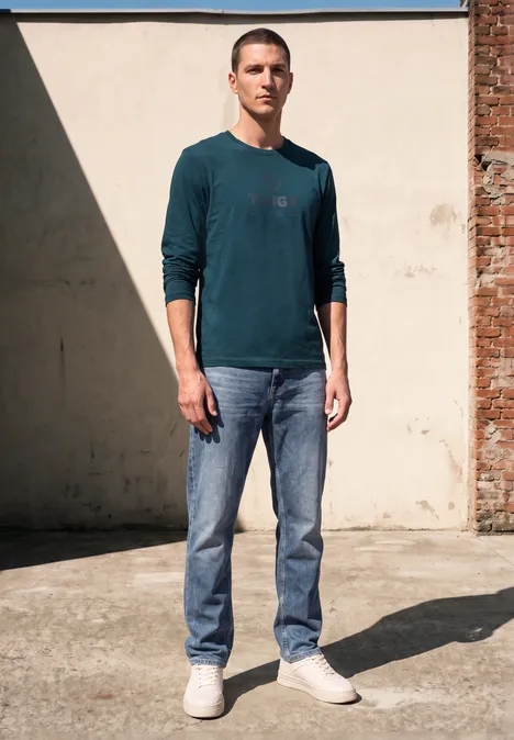 T-shirt à manches longues avec imprimé ton sur ton dark fjord blue Un homme en pull vert foncé et jean pose debout devant un mur, regard franc vers l’objectif.