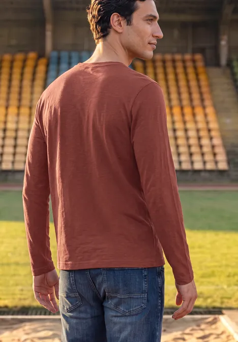 Longsleeve met tonale print raw copper red Ein Mann blickt von hinten auf das Spielfeld, trägt ein rostfarbenes Langarmshirt und Jeans.